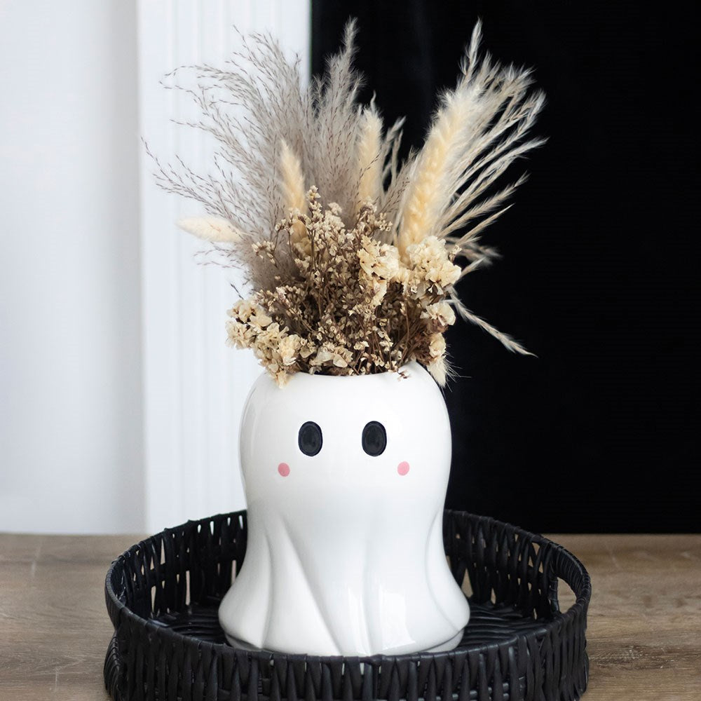 Ghost-shaped vase with dried flowers on a black tray against a white curtain background
