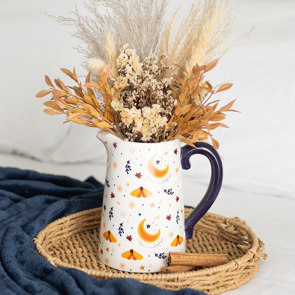 Decorative pitcher vase designed with moons, moths and falling leaves with dried flowers on a mat