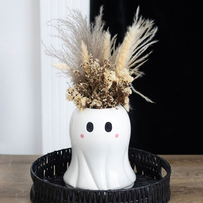 Ghost-shaped vase with dried flowers on a black tray against a white curtain background