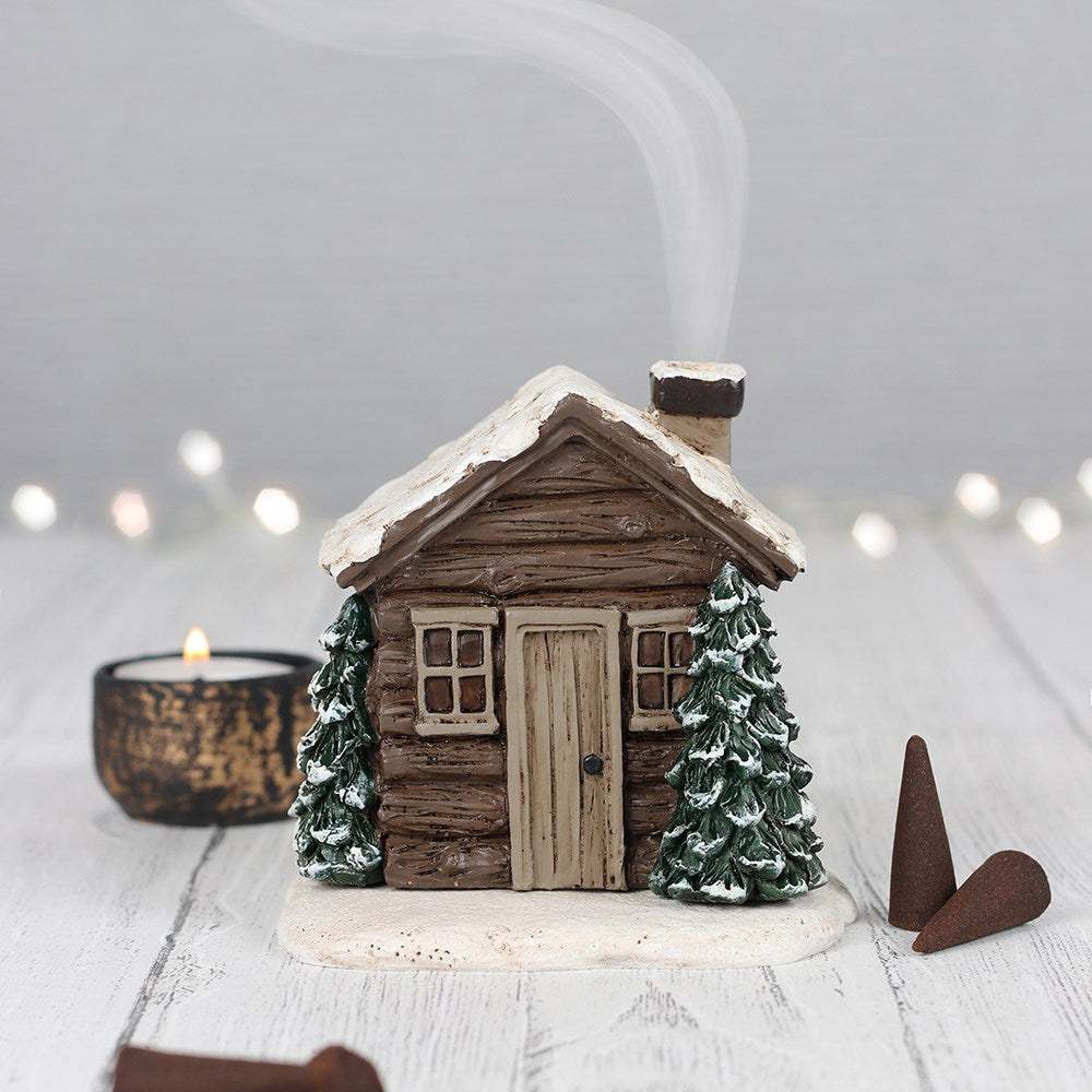 Dark brown log cabin incense cone holder, with snow on the roof and trees on the side. Smoke comes out of chimney. 