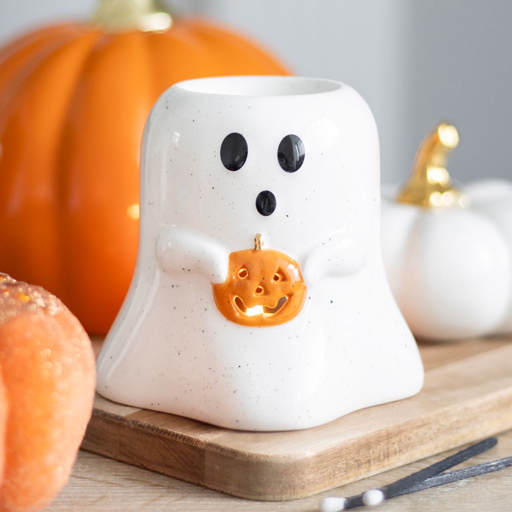 Ceramic ghost-shaped oil burner with a pumpkin design on a wooden surface with pumpkins in the background.