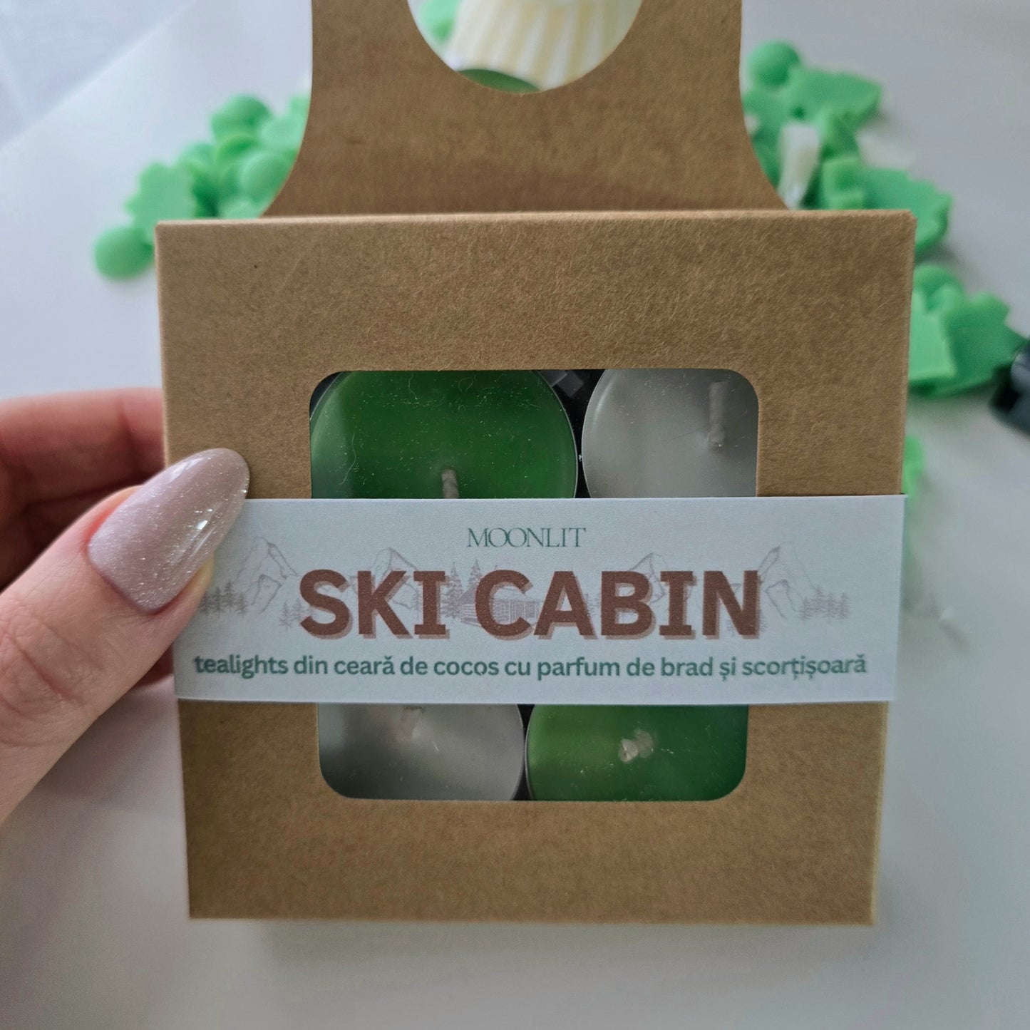 Box of 'Ski Cabin' tealights with a hand holding it, on a white background.