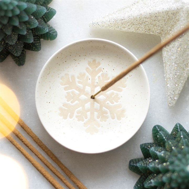 White ceramic dish with snowflake design holding an incense stick, surrounded by decorative elements on a light background.