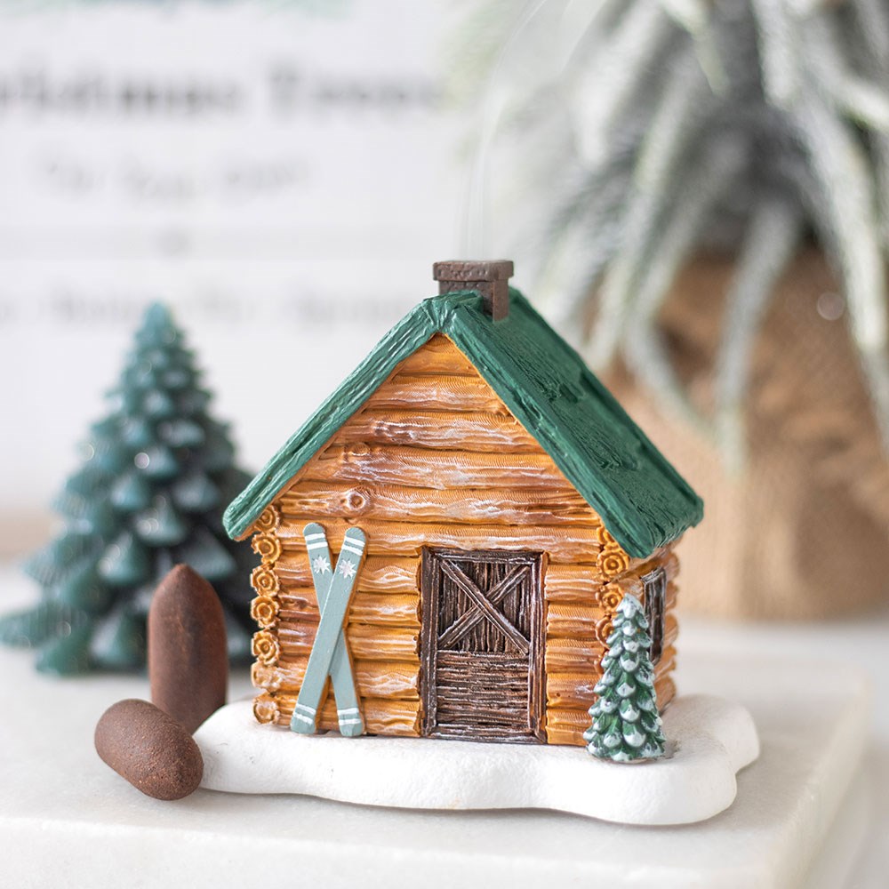 Winter Wonderland Ski Chalet Incense Burner - Miniature log cabin with green roof, skis, and small trees on a white like snowed surface.