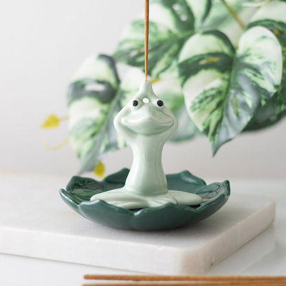 Green ceramic incense stick holder shaped like a pastel green frog that is in a yoga position on a dark green leaf.